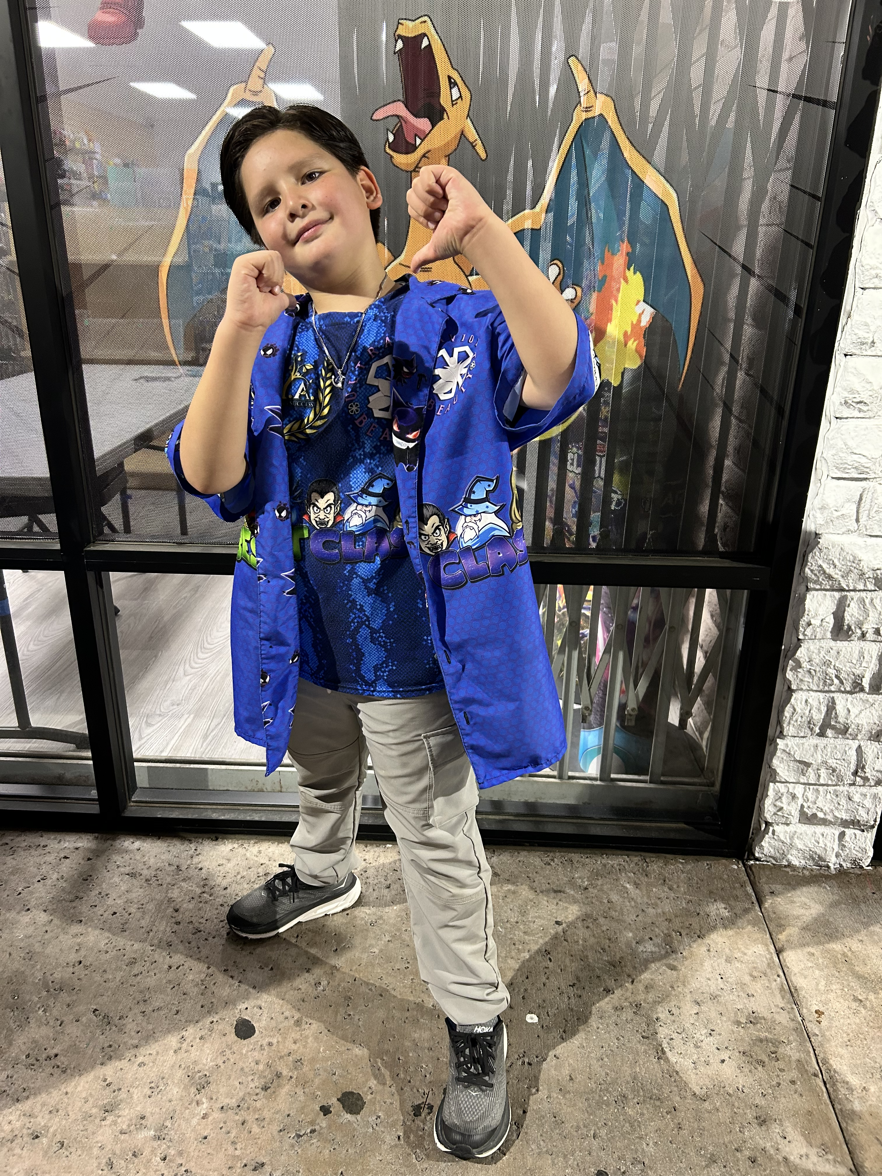 A confident child strikes a cool pose, giving playful thumbs-up and thumbs-down gestures while standing in front of a bold Charizard artwork. Dressed in a vibrant blue shirt with fun designs, they exude energy and charisma.